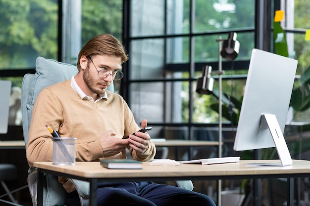 A young programmer freelancer sits at the desk in the office works at the computer holds a phone in