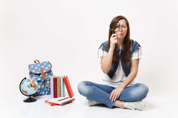 Young puzzled interested woman student holding looking on magnifying glass sitting near globe, backpack, school books isolated