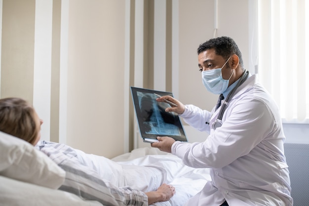 Young radiologist in whitecoat and protective mask commenting lung x-ray image of sick female patient while sitting by her bed in chamber