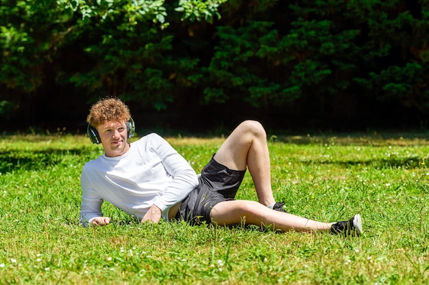 Young red-haired man in headphones lies on the green grass on a sunny day