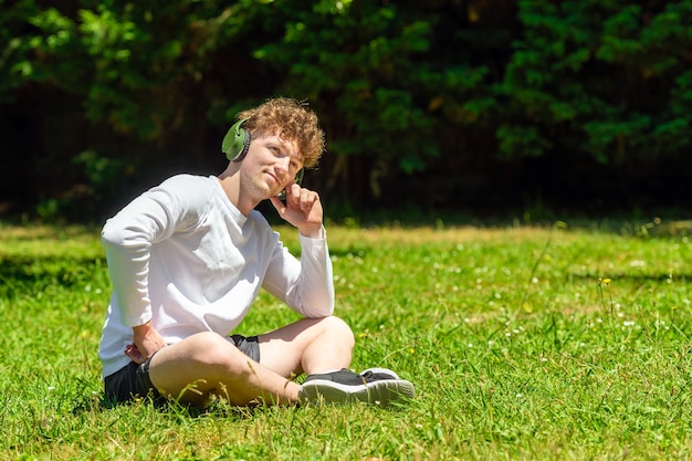 Young red-haired man with headphones sitting on the green grass on a sunny day