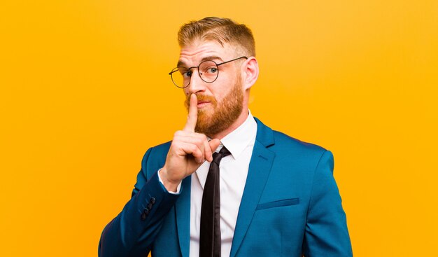 Young red head businessman asking for silence and quiet, gesturing with finger in front of mouth, saying shh or keeping a secret against orange 