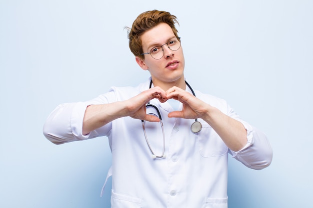 Young red head doctor smiling and feeling happy, cute, romantic and in love, making heart shape with both hands against blue wall