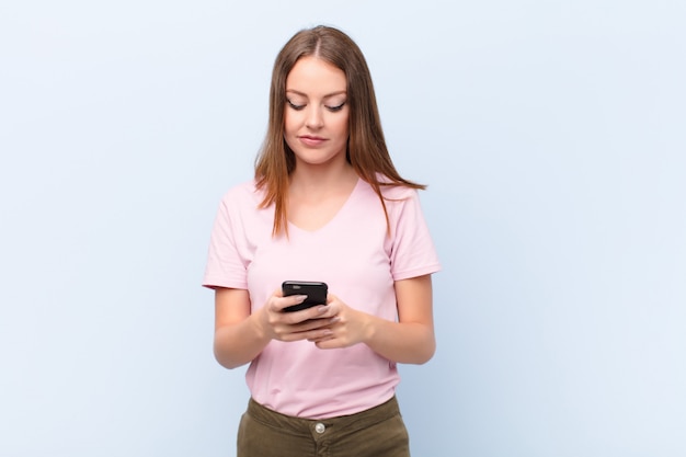 Young red head woman  against flat wall with a smart phone