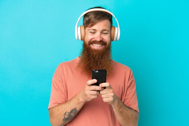 Young reddish caucasian man isolated on blue background listening music and looking to mobile