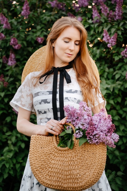 A young redhaired girl in a hat holds a wicker bag with blooming purple lilacs of greenery in spring