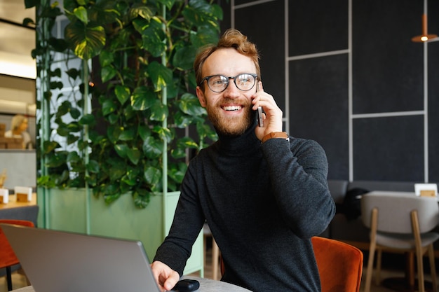 A young redhaired guy a programmer or entrepreneur in glasses sits in a stylish cafe at a laptop and speaks on the phone Freelancer works remotely Online communication Small business