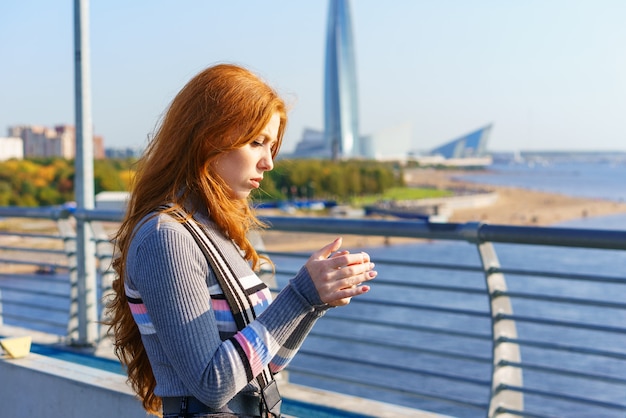 Young redhaired woman of caucasian ethnicity  with a phone in her hand stands on a bridge overlooking the city