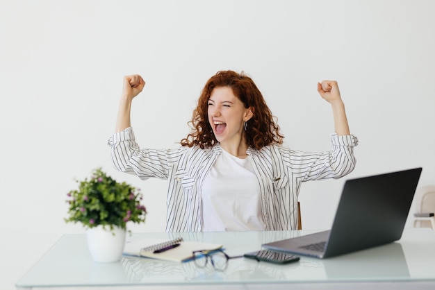 Young redhead curly woman working with her laptop raising fist after a victory winner concept