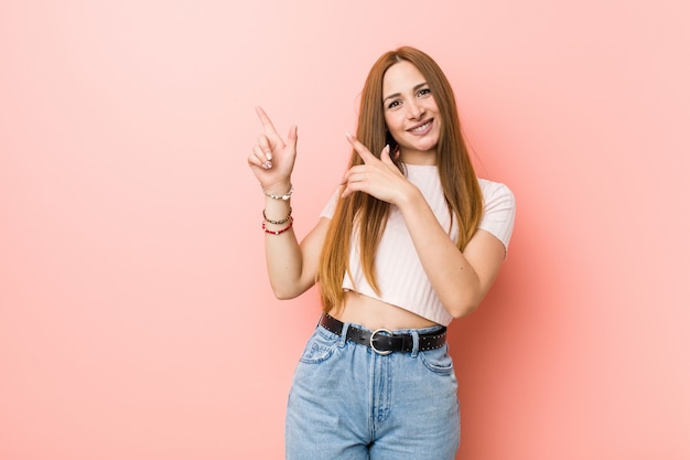 Young redhead ginger woman against a pink wall pointing with forefingers to a copy space, expressing excitement and desire.