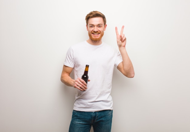 Young redhead man fun and happy doing a gesture of victory. Holding a beer.