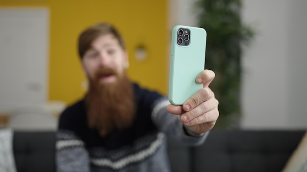 Young redhead man make selfie by smartphone sitting on sofa at home