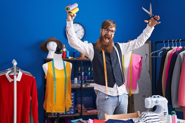 Young redhead man tailor smiling confident holding scissors and thread at clothing factory