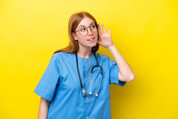 Young redhead nurse woman isolated on yellow background listening to something by putting hand on the ear