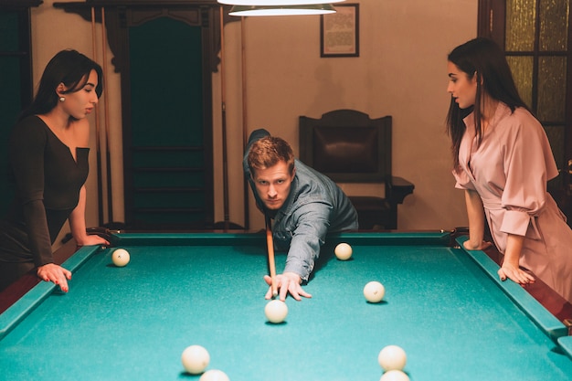 Young redhead player stand between two models. He aims into billiard ball. Young women stand and look at gamel. They lean to table.