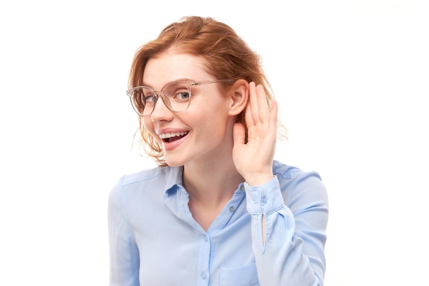 Young redhead woman in business shirt smiling and holding hand near ear listening carefully hearing intently isolated on white background