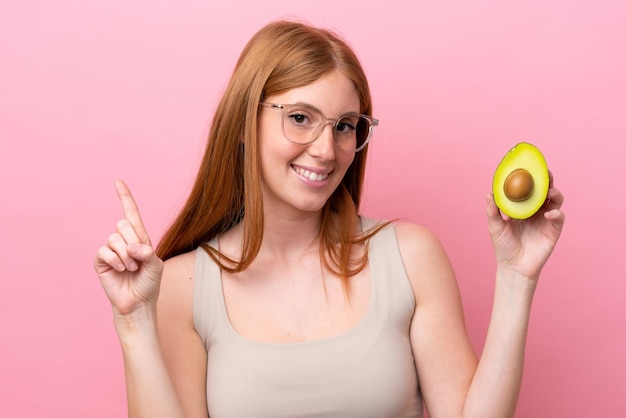 Young redhead woman holding an avocado isolated on pink background showing and lifting a finger in sign of the best