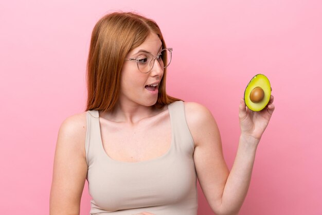 Young redhead woman holding an avocado isolated on pink background with surprise expression while looking side