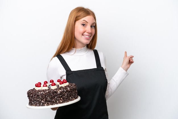 Young redhead woman holding birthday cake isolated on white background pointing back