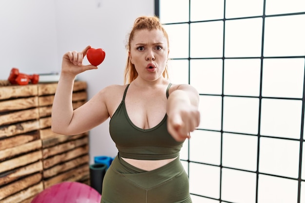 Young redhead woman holding heart at the gym pointing with finger to the camera and to you, confident gesture looking serious