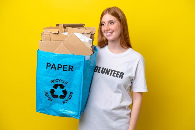 Young redhead woman holding a recycling bag full of paper to recycle isolated on yellow background looking to the side and smiling