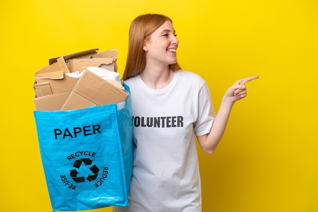 Young redhead woman holding a recycling bag full of paper to recycle isolated on yellow background pointing finger to the side and presenting a product