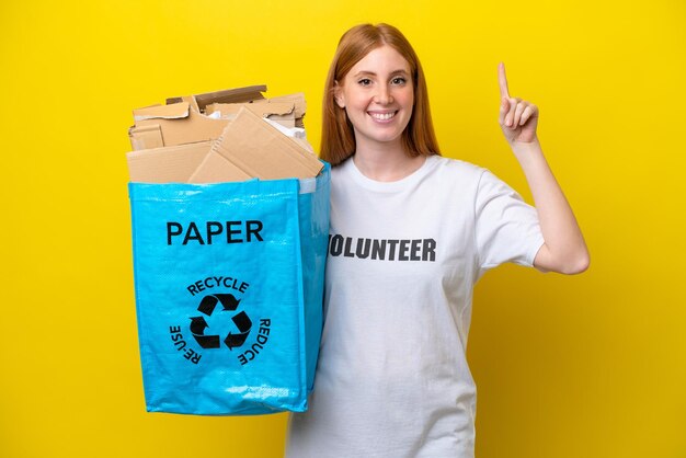 Young redhead woman holding a recycling bag full of paper to recycle isolated on yellow background pointing up a great idea