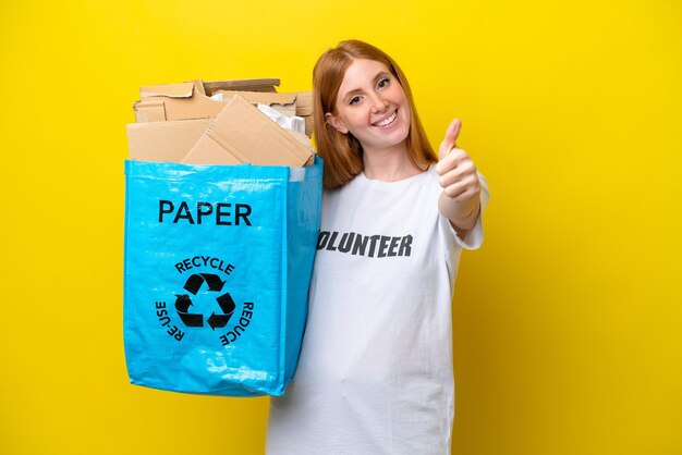 Young redhead woman holding a recycling bag full of paper to recycle isolated on yellow background with thumbs up because something good has happened