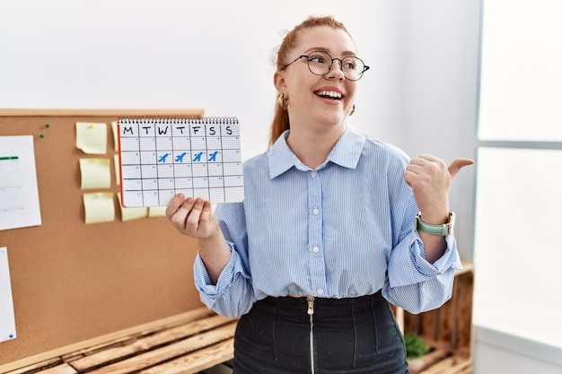 Young redhead woman holding travel calendar at the office pointing thumb up to the side smiling happy with open mouth