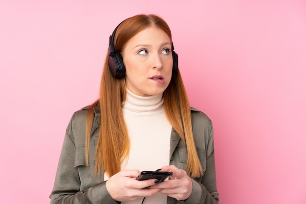 Young redhead woman over isolated pink wall listening music with a mobile and thinking