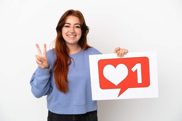 Young redhead woman isolated on white background holding a placard with Like icon and celebrating a victory