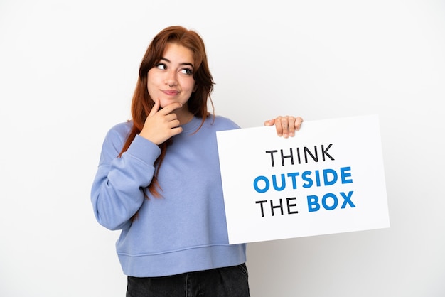 Young redhead woman isolated on white background holding a placard with text Think Outside The Box and thinking