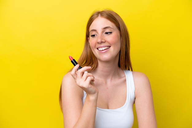 Young redhead woman isolated on yellow background holding red lipstick