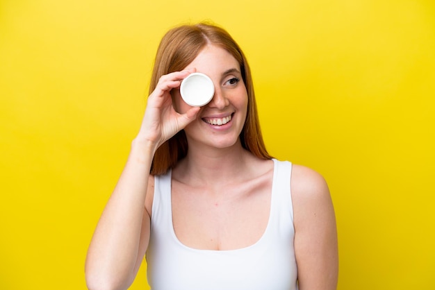 Young redhead woman isolated on yellow background with moisturizer and happiness