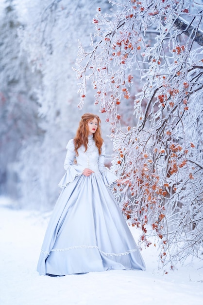 Young redhead woman a princess walks in a winter forest in a blue dress Frost and snow