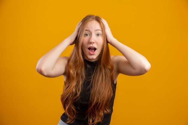 Young redhead woman raising hands to head, open-mouthed, feeling extremely lucky, surprised, excited and happy against yellow wall.