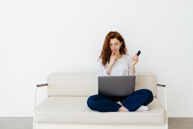 Young redhead woman relaxing on a sofa at home with her laptop on shops online with her credit card reading information