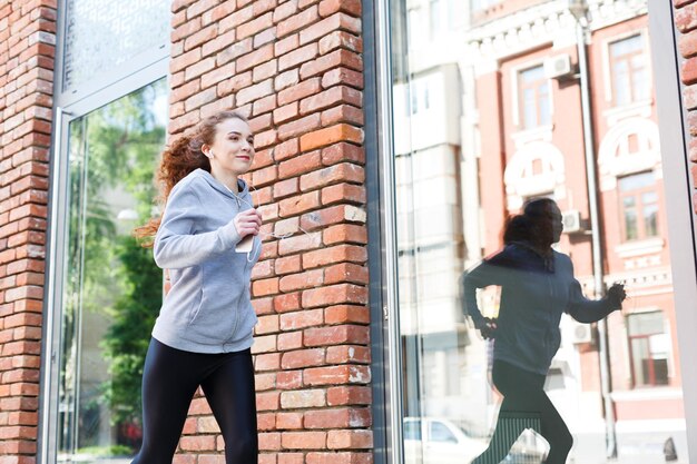 Young redhead woman running in city near brick building with mirror windows, copy space