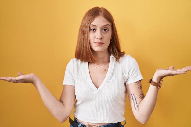 Young redhead woman standing over yellow background clueless and confused expression with arms and hands raised. doubt concept.