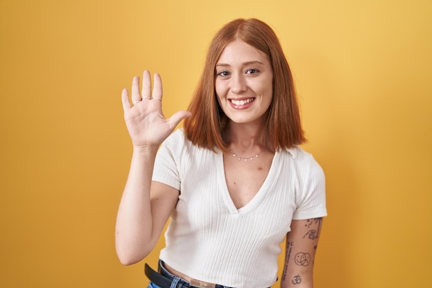 Young redhead woman standing over yellow background showing and pointing up with fingers number five while smiling confident and happy.