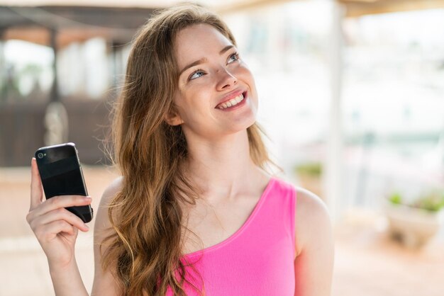 Young redhead woman using mobile phone at outdoors looking up while smiling