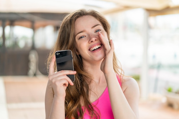 Young redhead woman using mobile phone at outdoors shouting with mouth wide open