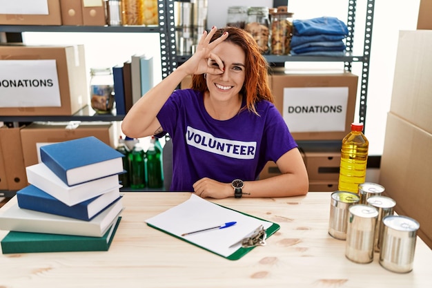 Young redhead woman wearing volunteer t shirt at donations stand doing ok gesture with hand smiling, eye looking through fingers with happy face.