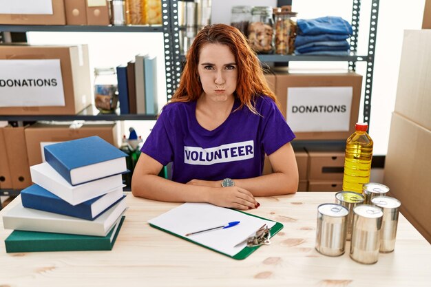 Young redhead woman wearing volunteer t shirt at donations stand puffing cheeks with funny face. mouth inflated with air, crazy expression.