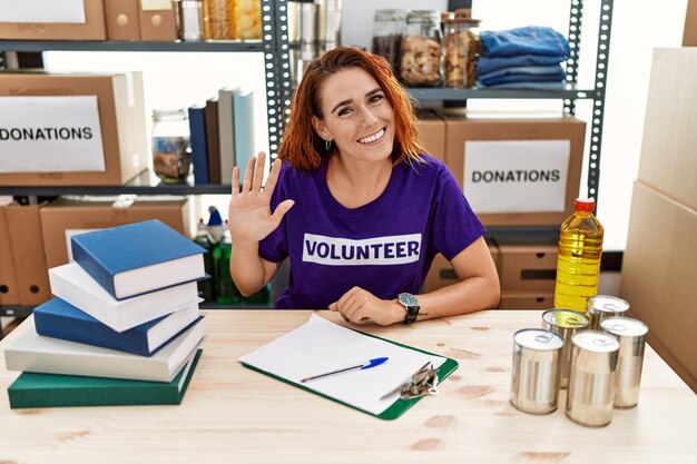 Young redhead woman wearing volunteer t shirt at donations stand waiving saying hello happy and smiling, friendly welcome gesture