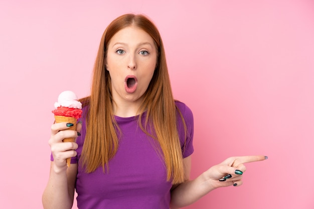 Young redhead woman with a cornet ice cream over isolated pink wall surprised and pointing side