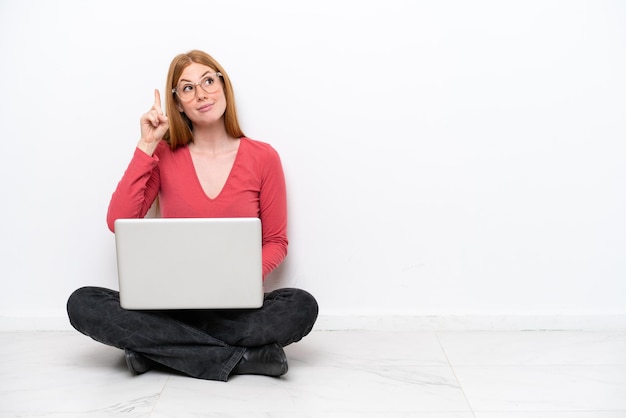 Young redhead woman with a laptop sitting on the floor isolated on white background intending to realizes the solution while lifting a finger up