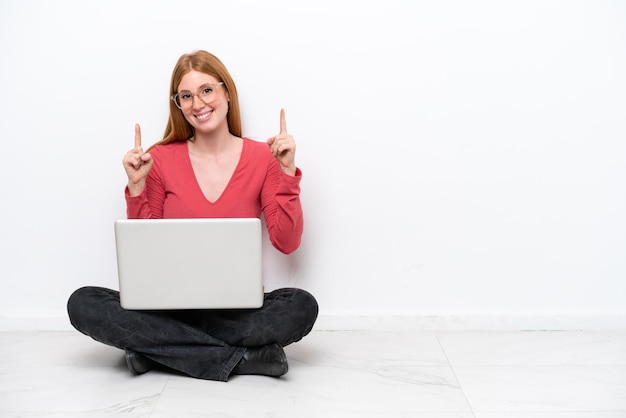 Young redhead woman with a laptop sitting on the floor isolated on white background pointing up a great idea