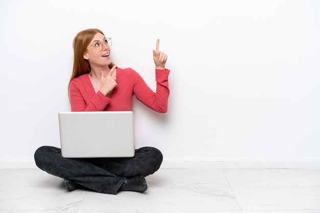 Young redhead woman with a laptop sitting on the floor isolated on white background pointing with the index finger a great idea