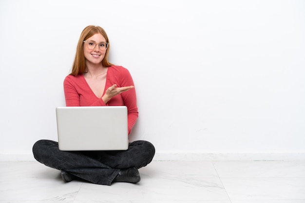 Young redhead woman with a laptop sitting on the floor isolated on white background presenting an idea while looking smiling towards